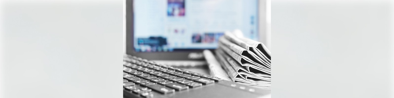 Eine Nahaufnahme einer Laptop-Tastatur mit einem Stapel gefalteter Zeitungen daneben und einem Computerbildschirm im Hintergrund.