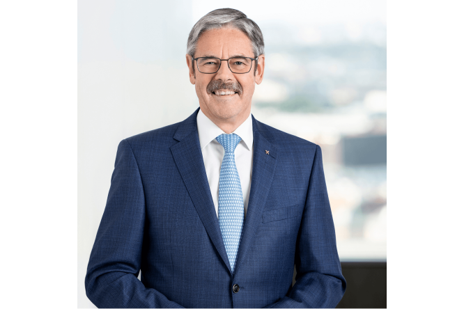 Obmann Mag. Erwin Hameseder, professionell gekleidet in einem blauen Anzug und einer hellblauen gemusterten Krawatte, steht in einem Innenraum. Im Hintergrund ist durch ein Fenster ein verschwommener Blick auf ein Büro oder eine Stadtlandschaft zu sehen. Die Szenerie deutet auf ein formelles Geschäftsumfeld hin.